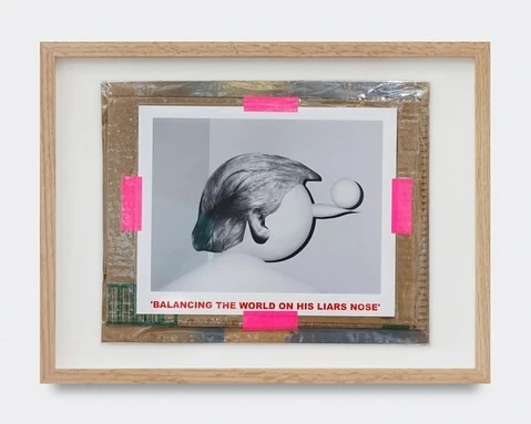 A framed photograph shows a sculptural head with gray hair balancing a white ball on its nose, secured by pink tape on cardboard backing.