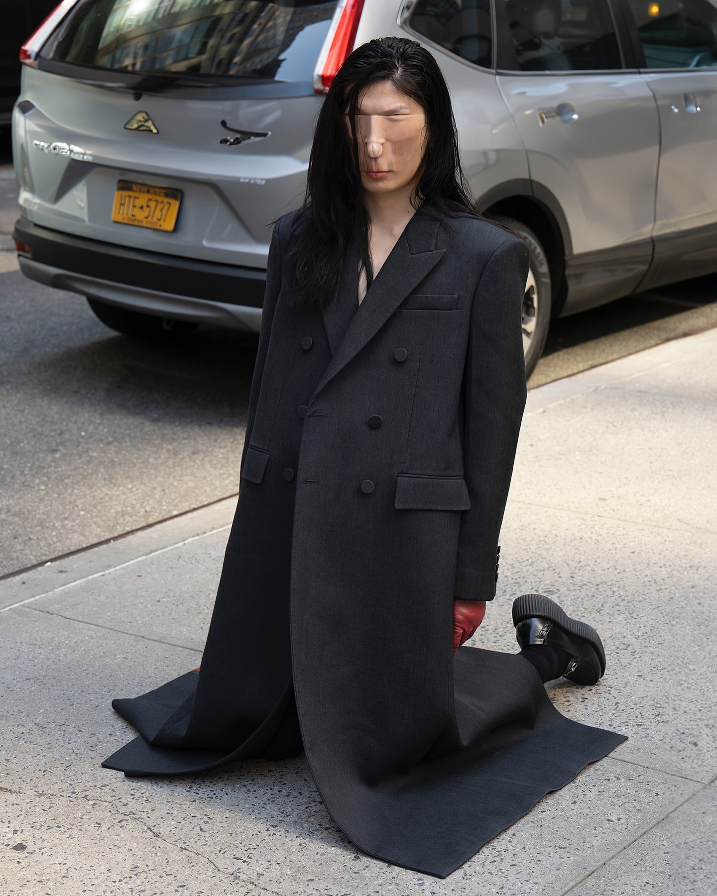 Person in oversized coat kneeling on sidewalk in urban setting with car in the background, showcasing unique fashion and urban lifestyle.