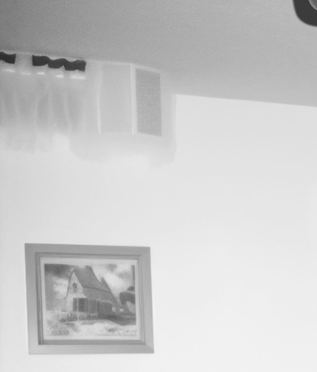 Interior wall with framed painting of a house, an air conditioning unit, and sheer curtains; grayscale, minimal decor.