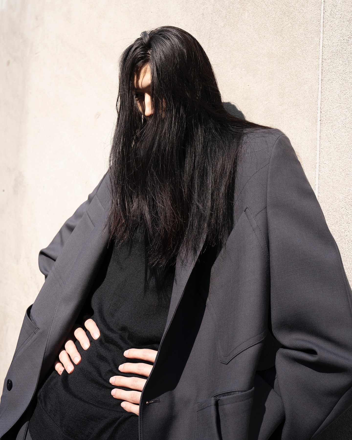 Model with long dark hair covering face wearing a charcoal grey blazer and black top leaning against a wall
