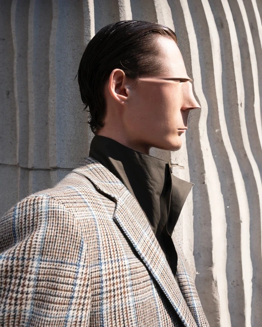 Adult male model in glen plaid blazer poses in an eye-level, medium shot next to a concrete wall with sunlight and shadow for fashion.