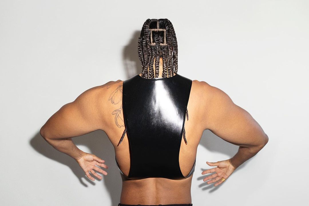 An adult person with braided hair is shot from the back, wearing a black leather-like harness, against a plain, light background.