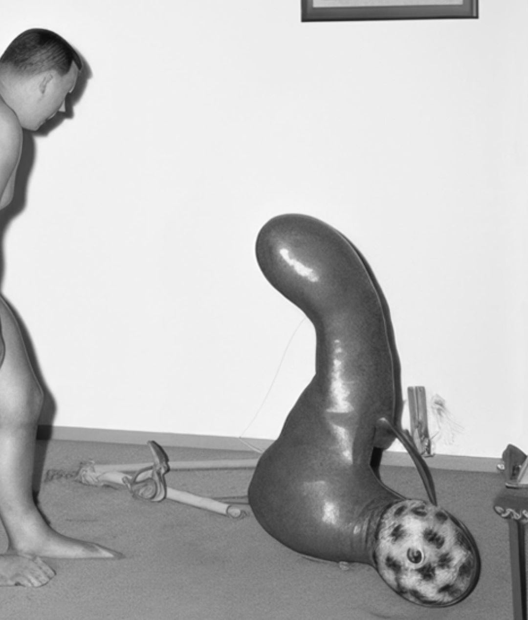 A shirtless man looks at a strange sculpture with a human-like head and a bulbous body in a white-walled indoor room in a black and white shot.
