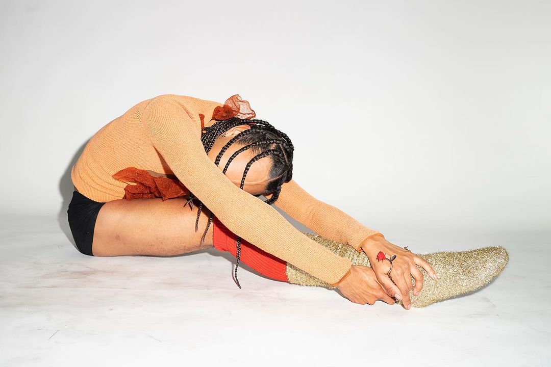 Person in golden socks and peach sweater stretches towards their feet in a white photo studio setting for fitness or fashion shoot.