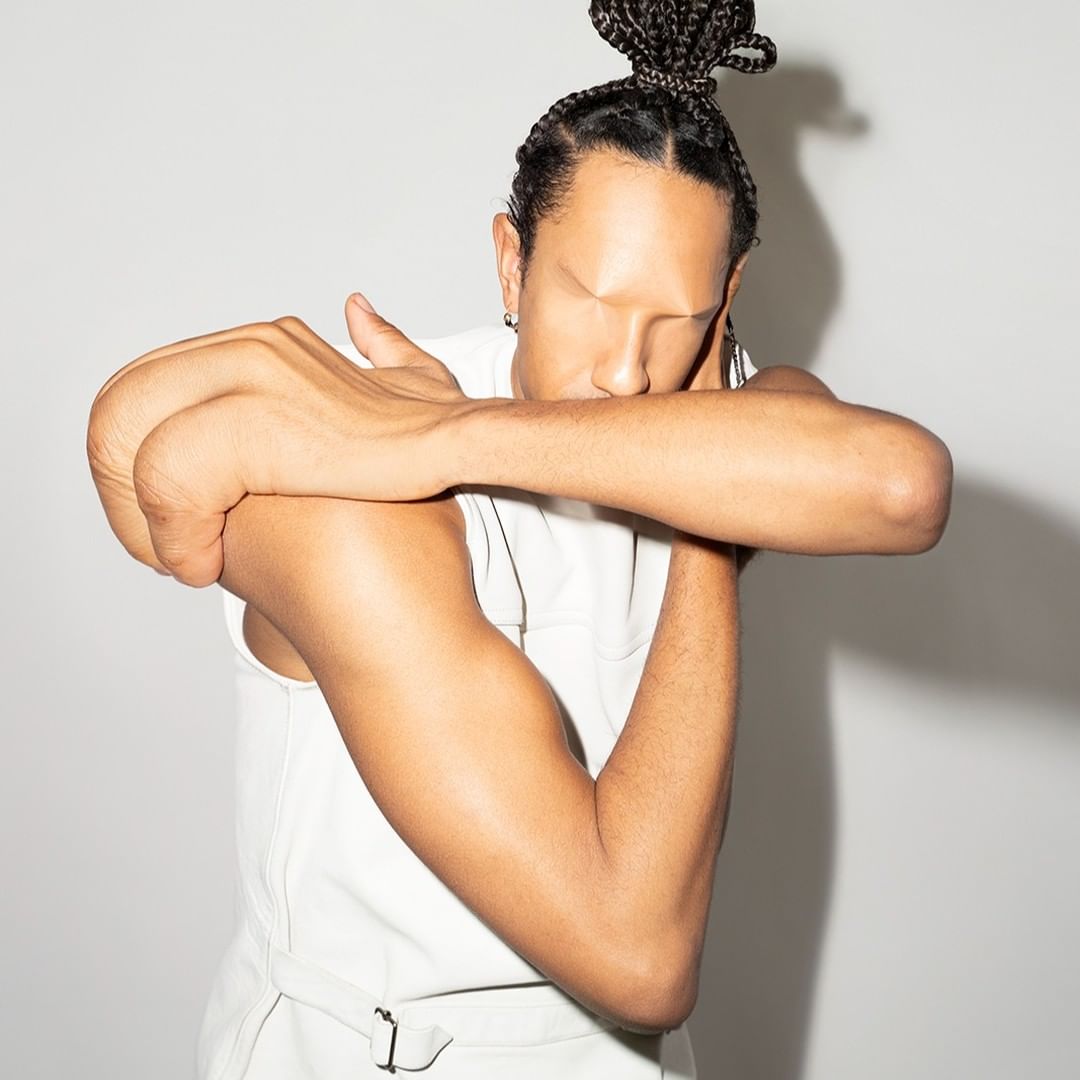 Adult male with braided hair posing in a sleeveless top in a studio. His face is partially hidden by his crossed arms against a grey backdrop.