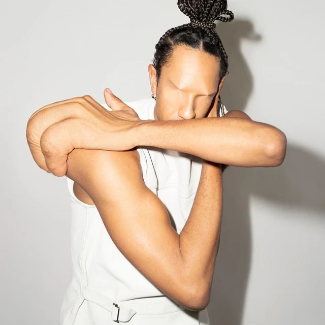 Adult male with braided hair posing in a sleeveless top in a studio. His face is partially hidden by his crossed arms against a grey backdrop.