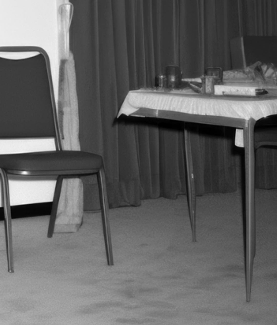 Black and white shot of a table with items on it, next to a chair in an indoor environment with a curtain backdrop.