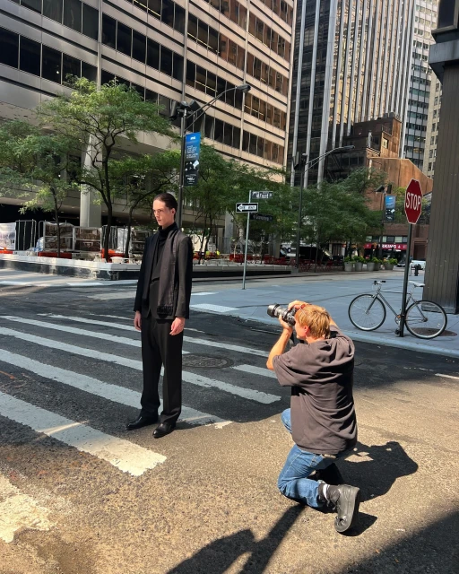 Male model poses on a crosswalk during an outdoor fashion shoot in a city with a photographer kneeling and shooting the model.