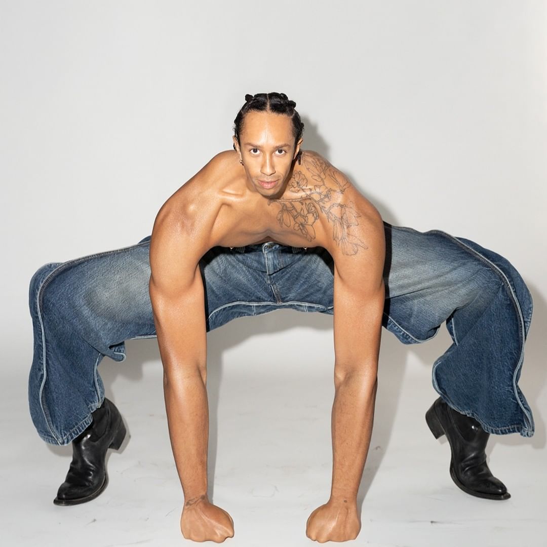 Adult male shirtless with tattoo, wearing blue jeans on all fours posing in a photo studio, black cowboy boots, and white background.
