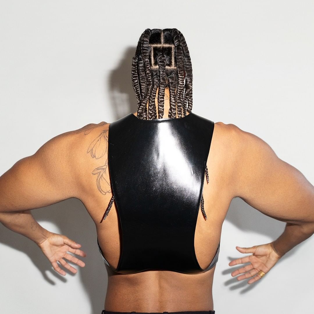 A person with braided hair faces away from the camera, wearing a shiny black top and presenting their palms in a studio setting.