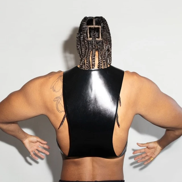 A person with braided hair faces away from the camera, wearing a shiny black top and presenting their palms in a studio setting.