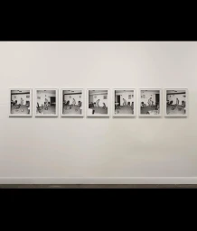 Seven framed black and white photographs displayed in a horizontal row on a plain white gallery wall