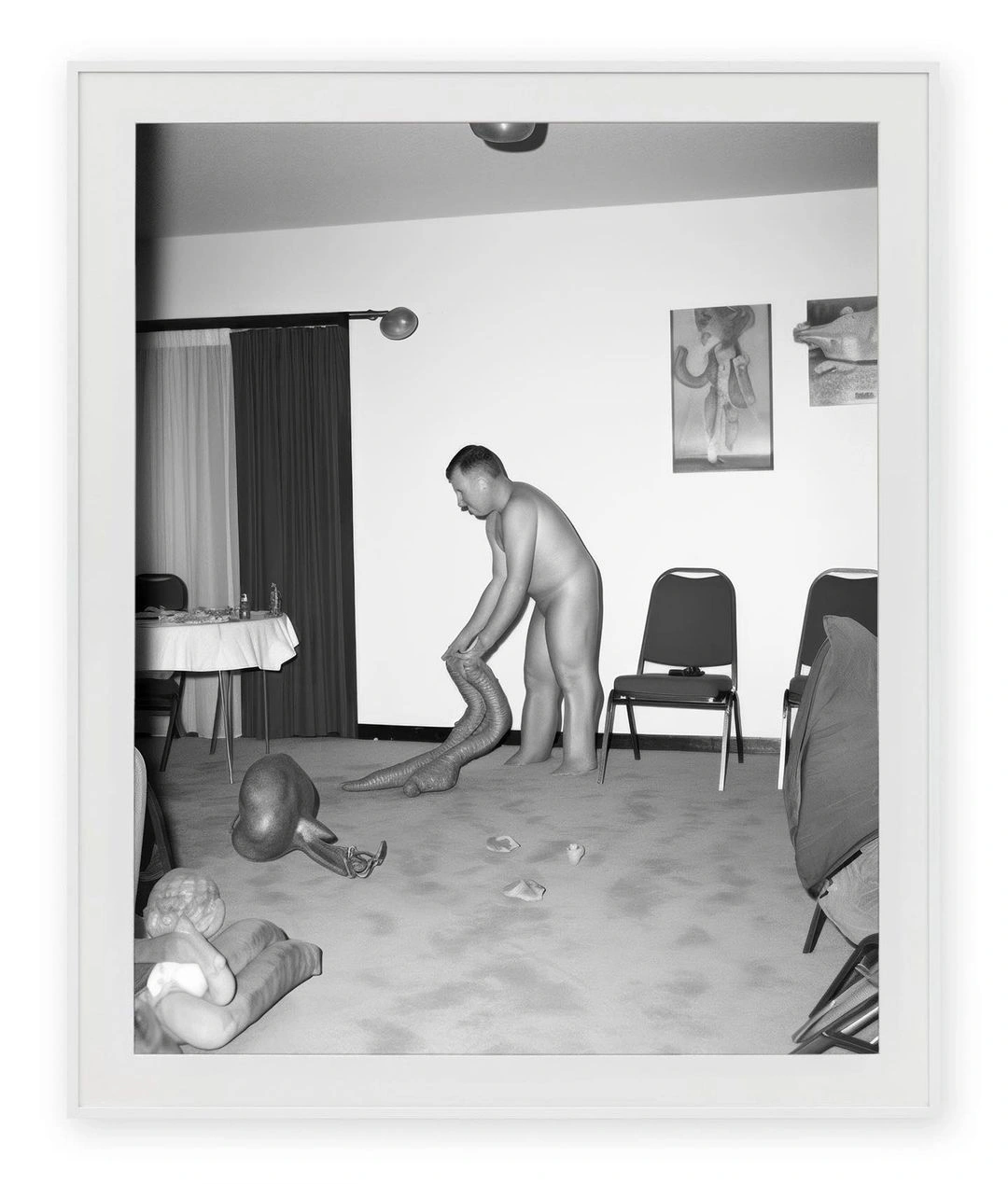 Black and white photograph of a man bending over in an interior room with chairs, a table, and surreal distorted body parts on the floor