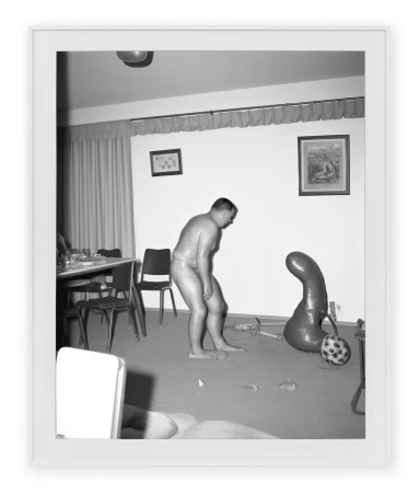 Monochrome photograph by Asger Carlsen of a nude man standing in a living room beside an abstract form