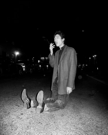 Man sitting on asphalt at night wearing an oversized jacket and smoking a cigarette, photographed by Asger Carlsen.
