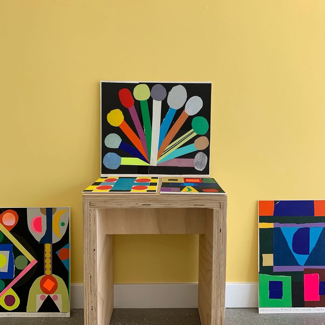 Arrangement of vibrant geometric abstract paintings and artworks displayed on a light wood table against a pale yellow wall.