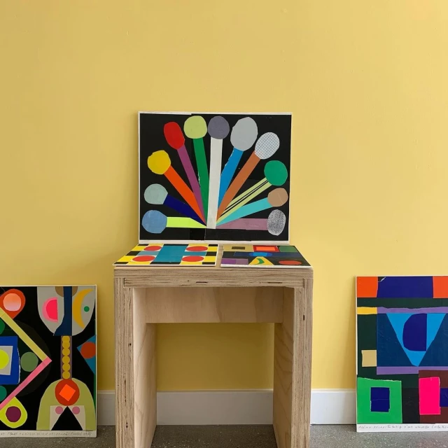 Arrangement of vibrant geometric abstract paintings and artworks displayed on a light wood table against a pale yellow wall.