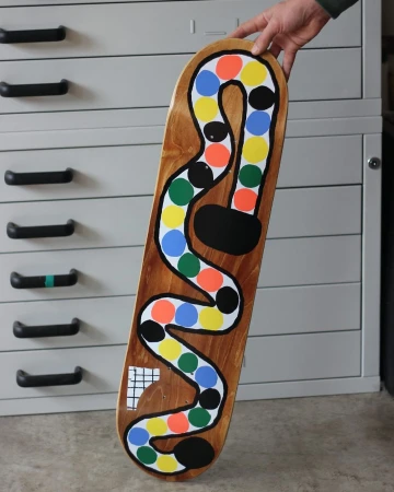 Close-up of a colorful custom skateboard held by a hand with a drawing of a winding path of colorful dots, placed in front of a filing cabinet.