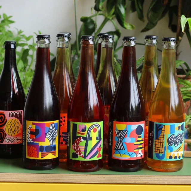 Nine bottles of GALLIA beer with colorful abstract labels are arranged on a yellow surface, set against a background of green foliage.