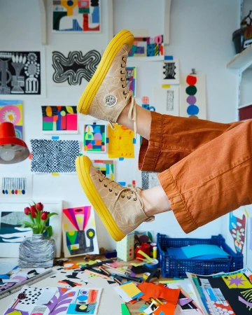 Elevated shot of brown high-top sneakers with yellow soles, styled with tan slacks, set against an artist studio backdrop with artwork.
