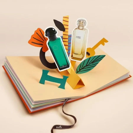 Two perfume bottles on a book with paper art accents. One bottle is green, the other is clear, in a beauty or fragrance advertisement.