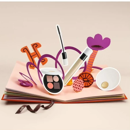 Cosmetic products arranged on a pop-up book, featuring eye shadow, mascara, lip gloss, and stylized paper cut-out decorations.