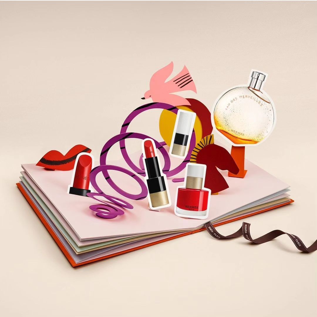 Pop-up book featuring perfume, lipstick, and nail polish bottles in a studio shot with a bird cutout and purple geometric figures.