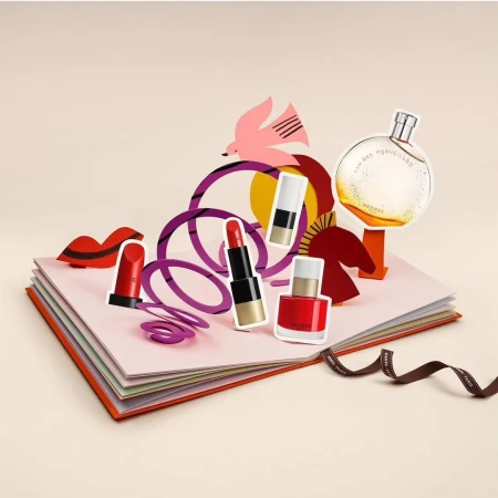 Pop-up book featuring perfume, lipstick, and nail polish bottles in a studio shot with a bird cutout and purple geometric figures.