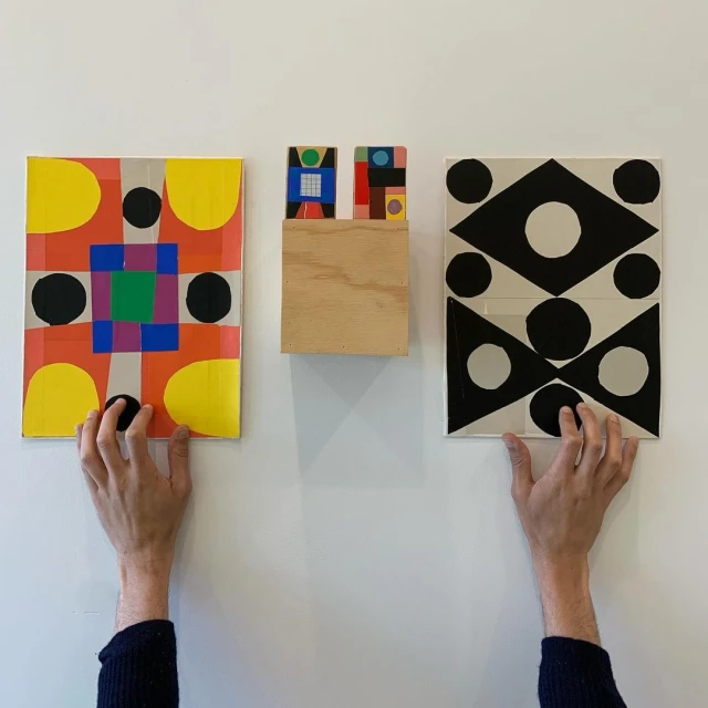 Two hands holding up colorful abstract geometric paintings against a white wall; bright and visually striking modern art display.