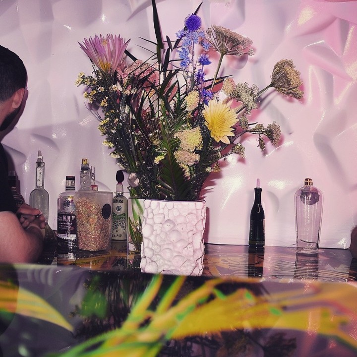 Indoor bar scene with a colorful floral arrangement in a textured vase, liquor bottles, and a glimpse of a person.