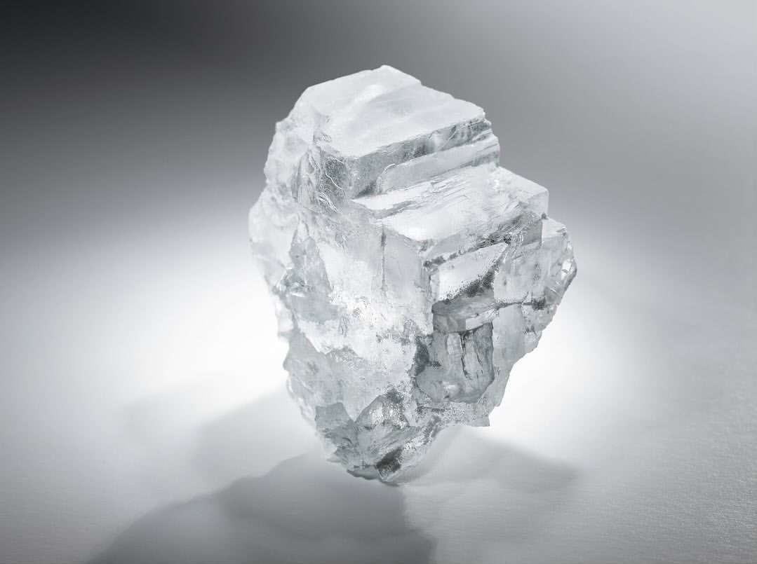 Close-up studio shot of a crystalline mineral specimen on a gray gradient background with a cast shadow showing its cubic structure.