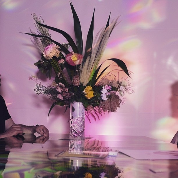 Elegant floral arrangement with mixed flowers and greenery in a crystal vase, set on a reflective table, soft pink lighting in the background.