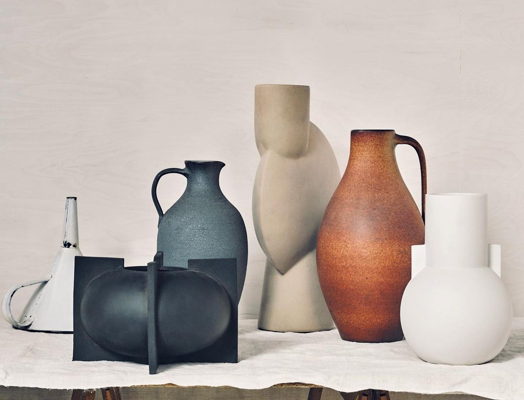 Still life photo featuring a variety of vases in neutral tones and varying shapes against a light, neutral backdrop on a linen tablecloth.