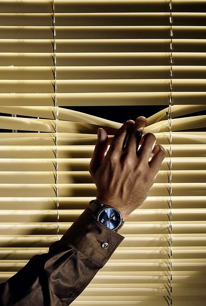 Close-up of an adult male hand opening a venetian blind to peek through it. The person is wearing a watch and brown shirt.