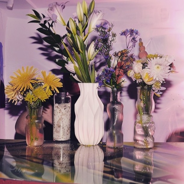 Still life of white lilies, yellow daisies and other colorful flowers in glass vases and white ceramic vase on reflective surface.