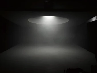Dark moody studio interior illuminated by a single circular overhead light casting a soft glow through haze