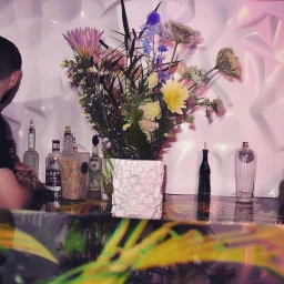Large floral arrangement in a textured white pot on a reflective bar counter with bottles and a person sitting