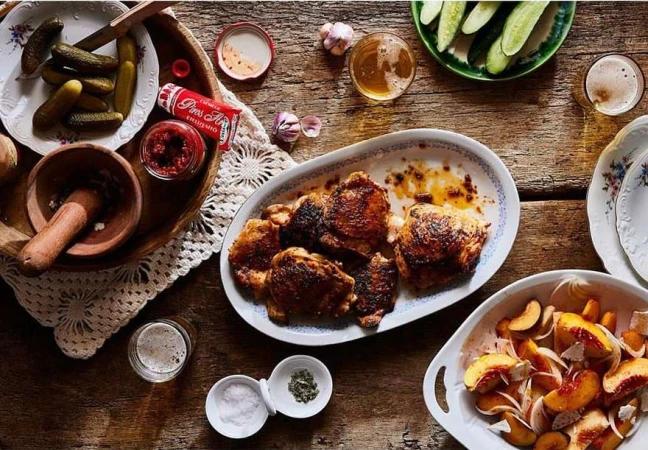 Overhead shot of a rustic wooden table with plates of grilled chicken, pickles, and peaches with onions, creating a rich food scene.