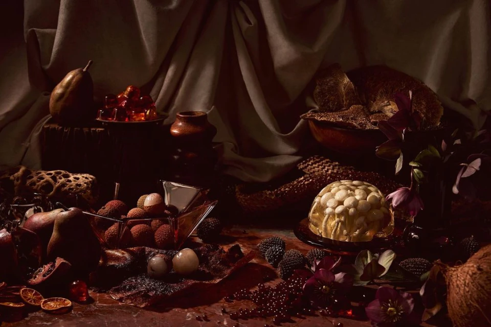 Dark still life photography featuring fruits like lychees and pears, along with flowers, set against draped fabric and a dark backdrop.