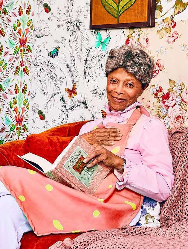 Elderly woman in pink apron and pink shirt is reading a book. She is sitting on a red couch with floral wallpaper behind her.