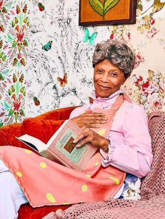 Elderly woman in pink apron and pink shirt is reading a book. She is sitting on a red couch with floral wallpaper behind her.