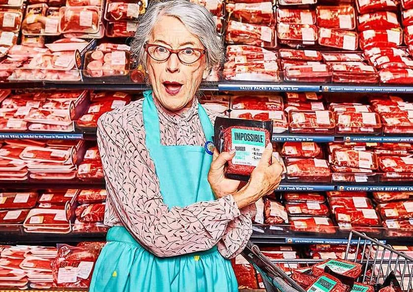 Elderly woman in a grocery store holds a package of Impossible Burger plant-based meat in front of a meat display case.