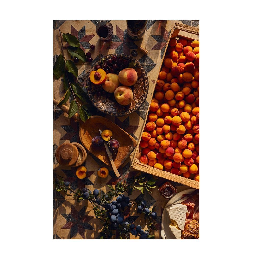 Overhead shot of peaches, apricots, plums, and cherries arranged on a patterned tile table with assorted serving dishes and utensils.