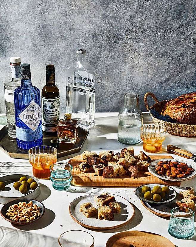Still life featuring liquor bottles, glasses, assorted olives, bread cubes, nuts, and almonds arranged on a white table against a textured background.