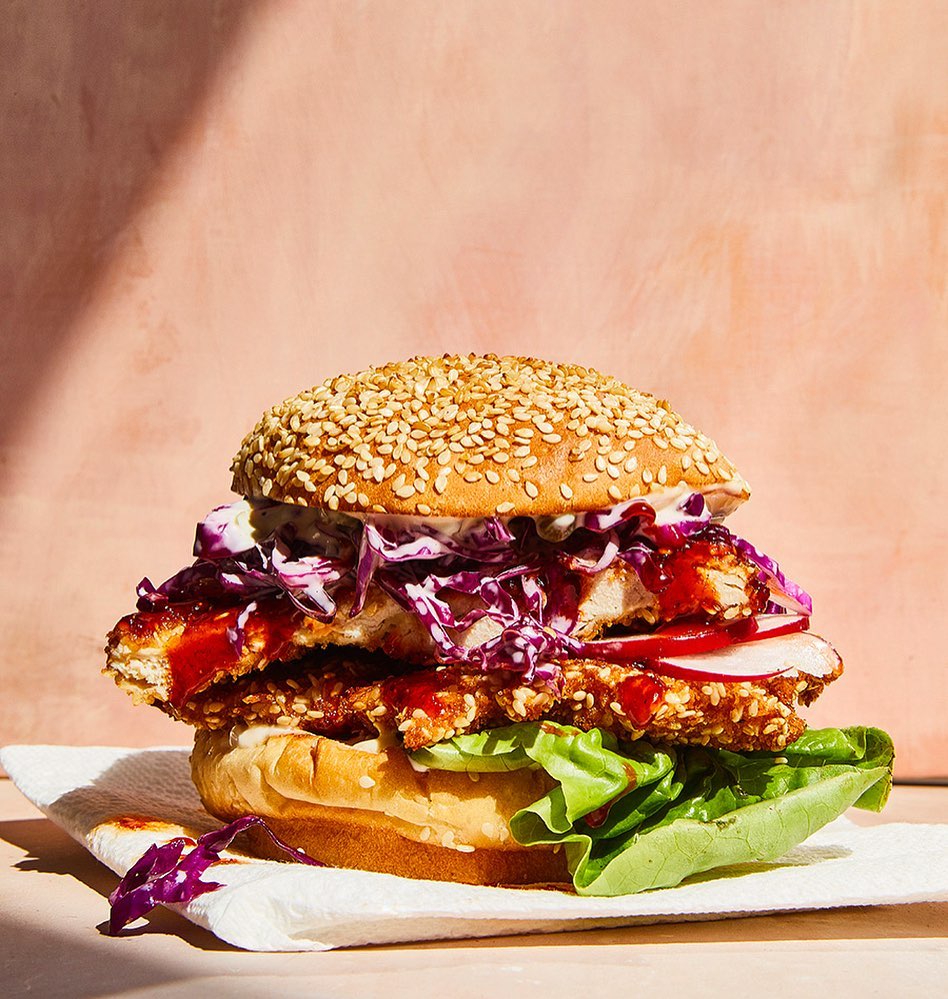 A towering sesame seed bun chicken sandwich with vibrant red cabbage slaw and crisp lettuce sits on a white napkin in a studio.