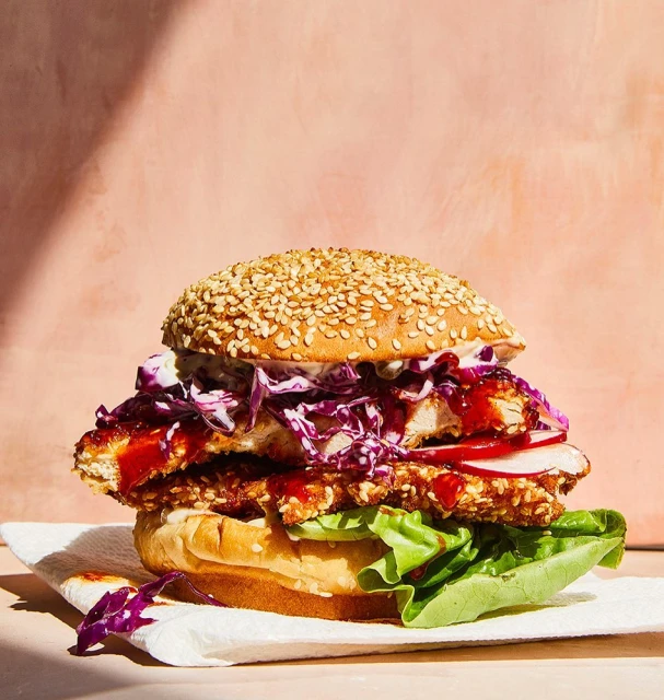 A towering sesame seed bun chicken sandwich with vibrant red cabbage slaw and crisp lettuce sits on a white napkin in a studio.