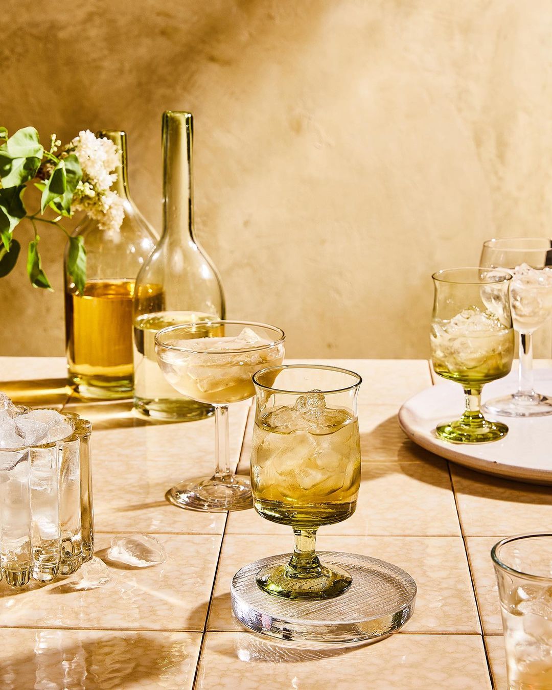 Still life captures a collection of clear glassware filled with golden liquid and ice, set on a tiled surface in warm lighting.