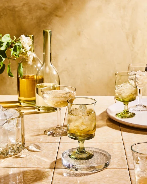 Still life captures a collection of clear glassware filled with golden liquid and ice, set on a tiled surface in warm lighting.