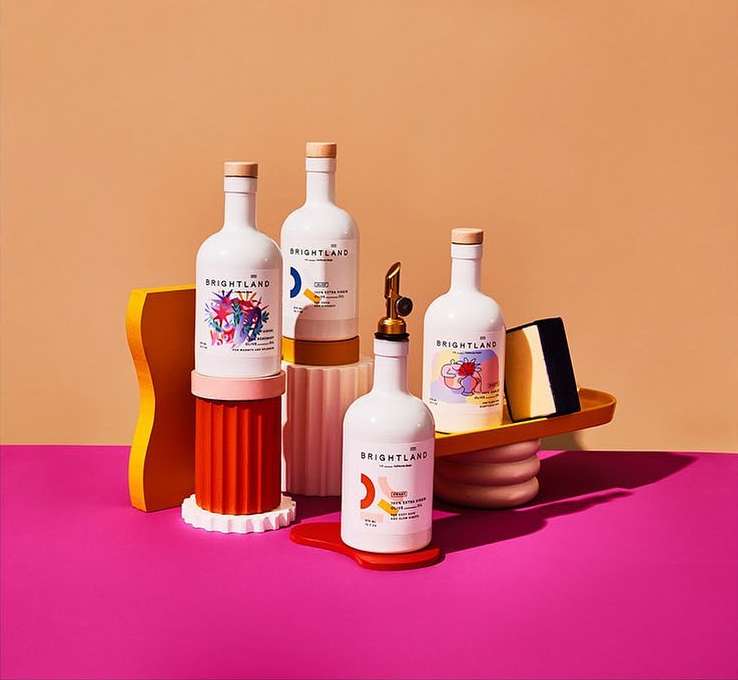 Five Brightland olive oil bottles with colorful geometric and abstract designs on a pink and peach background, product photography.