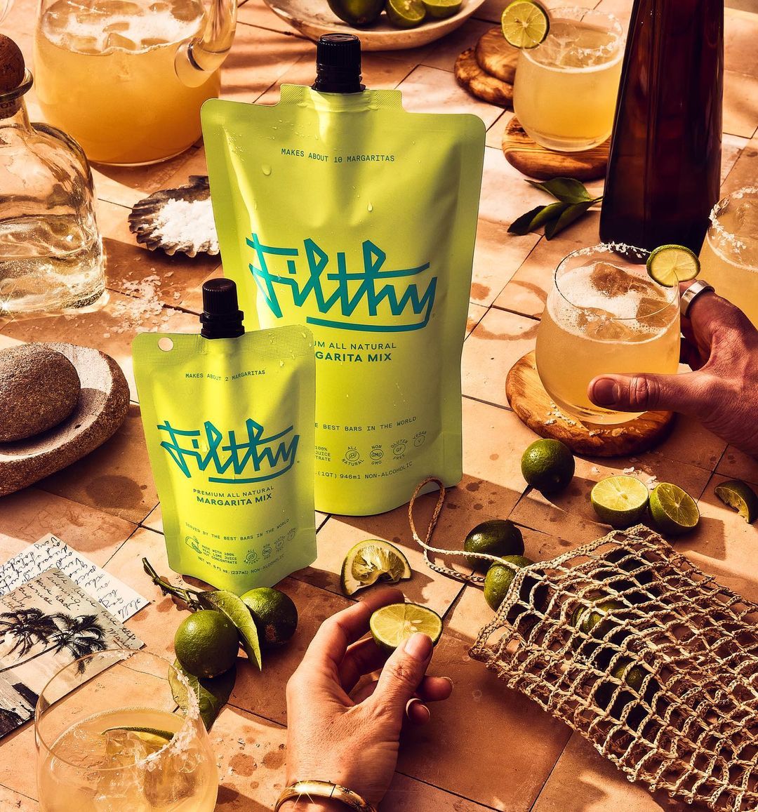 Two sizes of Filthy Margarita Mix stand on a tile surface with limes, margarita glasses, and hands reaching for the drinks.